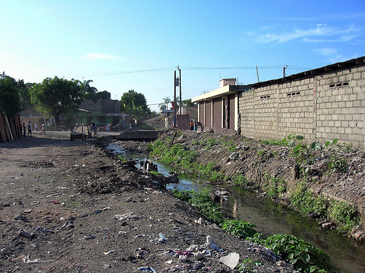 Absence d'assainissement à Cap-Haïtien, Haïti : les déchets à même le sol (incluant des sacs plastiques remplis d'excréments) bloquent les canaux de drainage qui débordent à la moindre pluie, et endommagent les infrastructures à côté (routes, bâtiments…).
