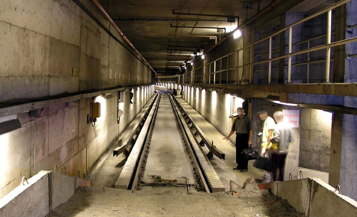 Vue depuis un butoir d’une voie sur pneu en coupe. On voit bien les barres de guidage et d’alimentation, les pistes de roulement et la voie standard, au milieu. Les personnages donnent l’échelle.